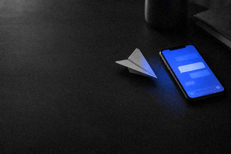 Бумажный самолётик рядом со смартфоном, экран светится синим чатом
